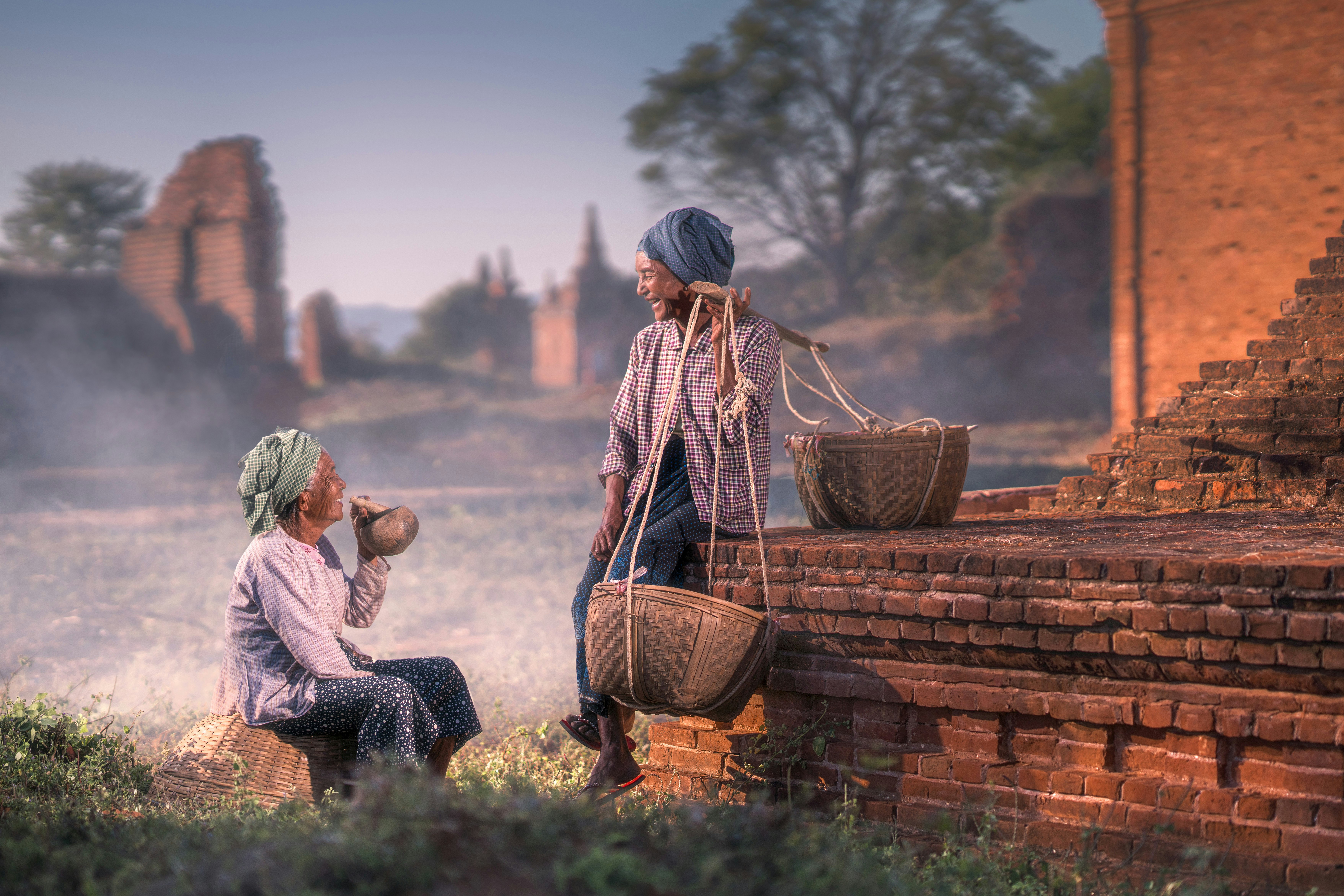 sitting person smiling beside man sitting on brown concrete pavement while carrying baskets on his left shoulder